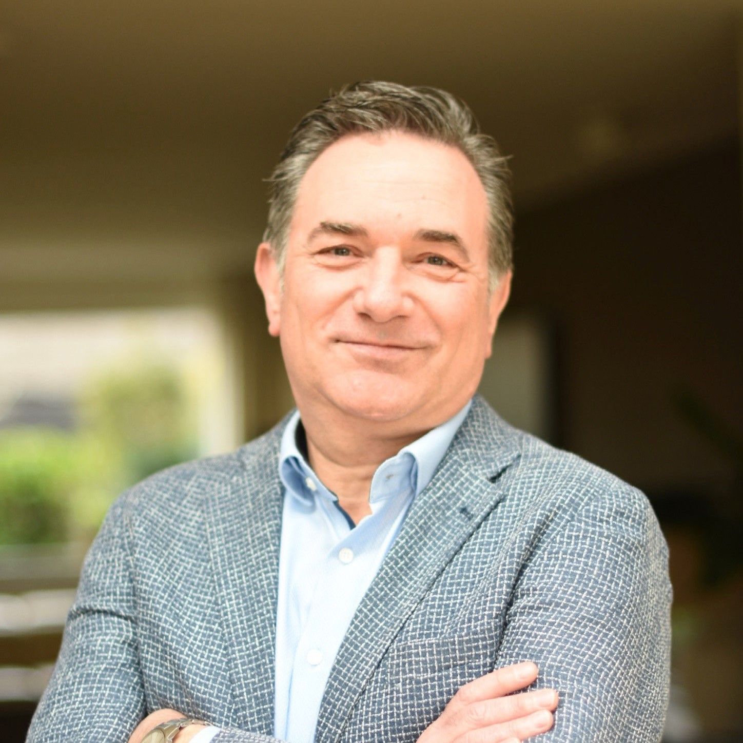 Smiling man in a light blue shirt and gray blazer, standing with arms crossed outdoors.