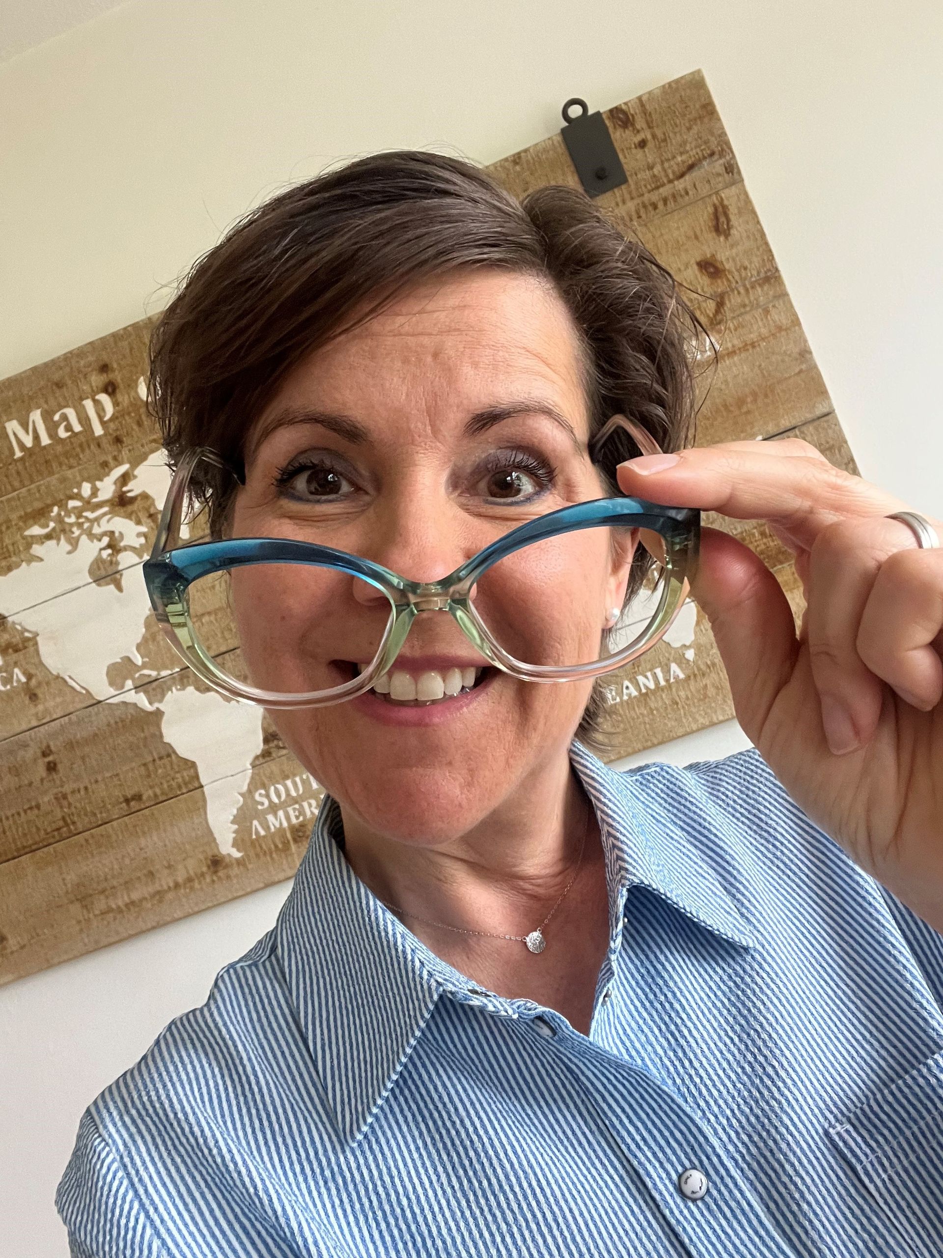 Smiling person holding turquoise eyeglasses in front of a map wall backdrop