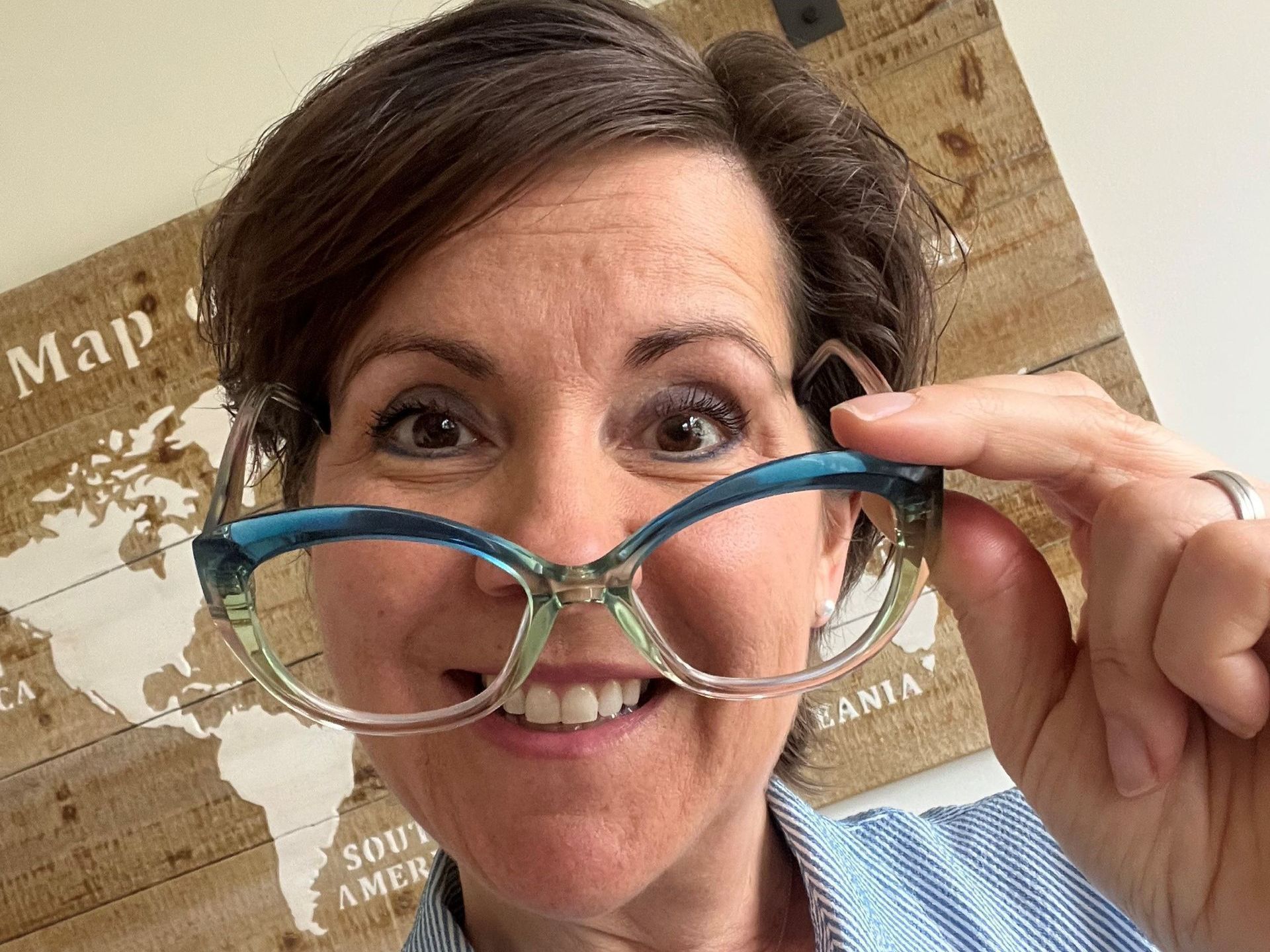 Woman holding blue eyeglasses up to her face, smiling in front of a map background