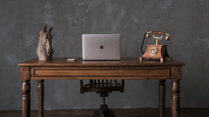 A wooden desk with a laptop, decorative sculpture, and vintage rotary phone against a gray wall.