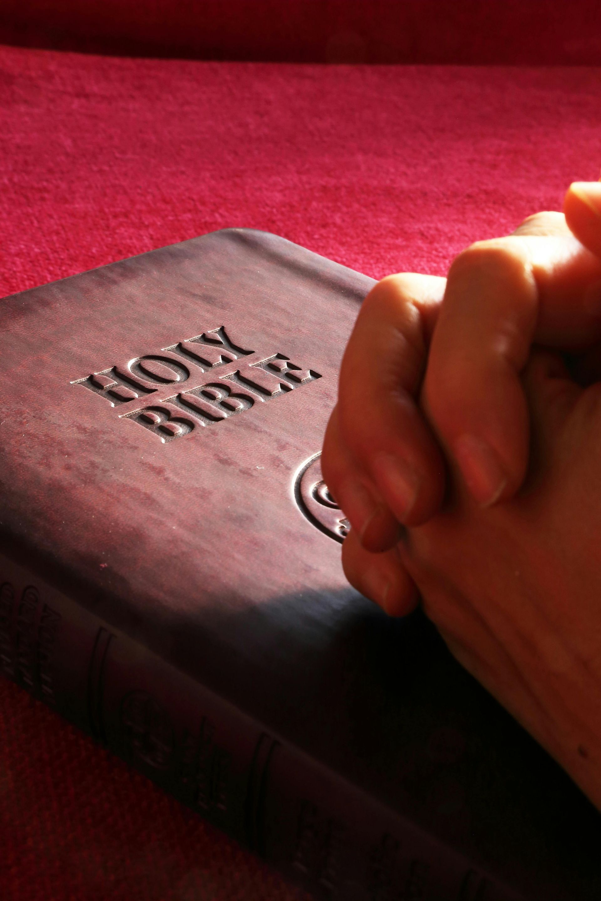 Hands clasped in prayer beside a Bible on a red surface.