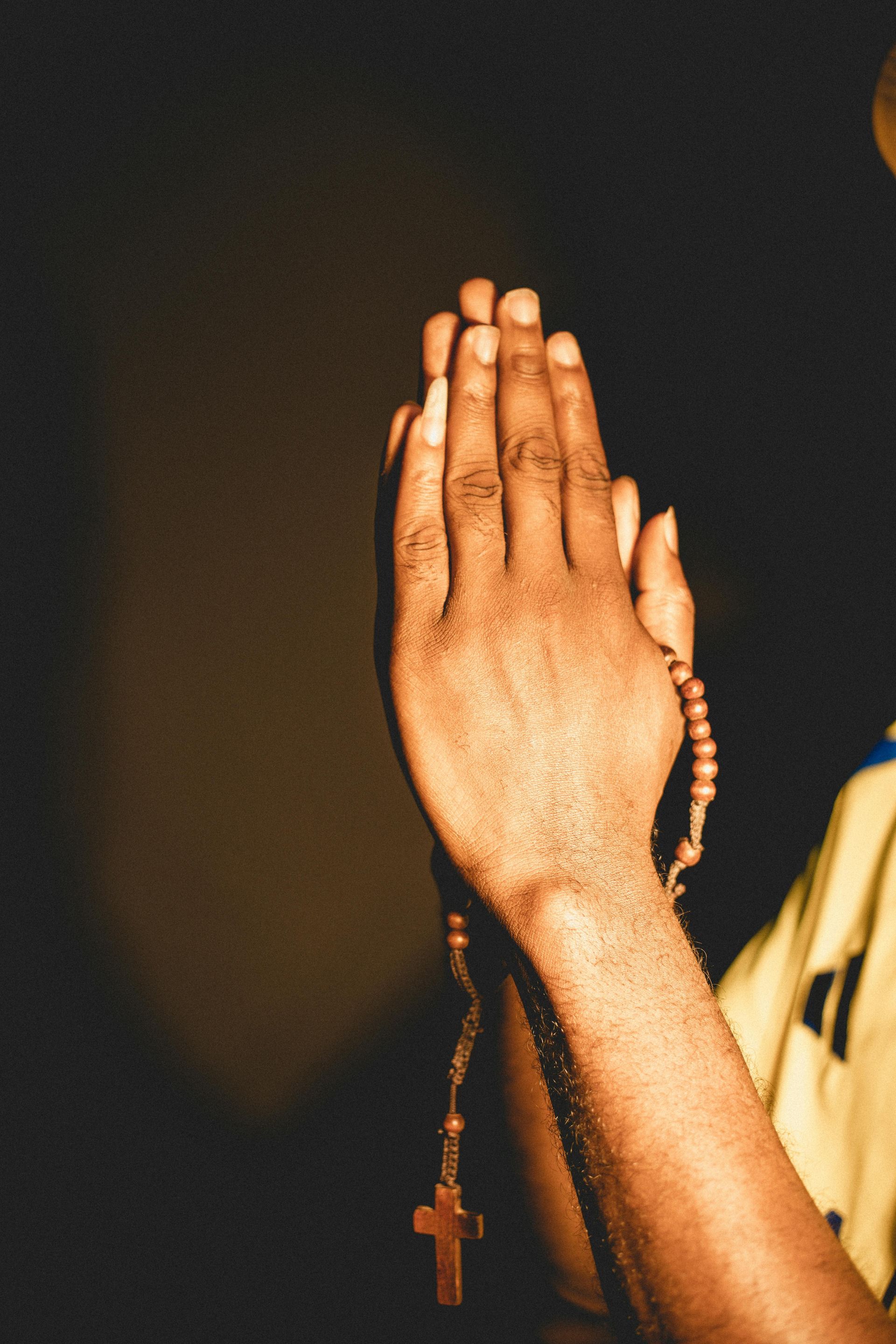 Hands clasped in prayer holding a rosary with a cross against a dark background