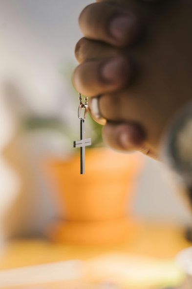 Hand holding a small cross pendant on a chain, with a blurred warm-toned background
