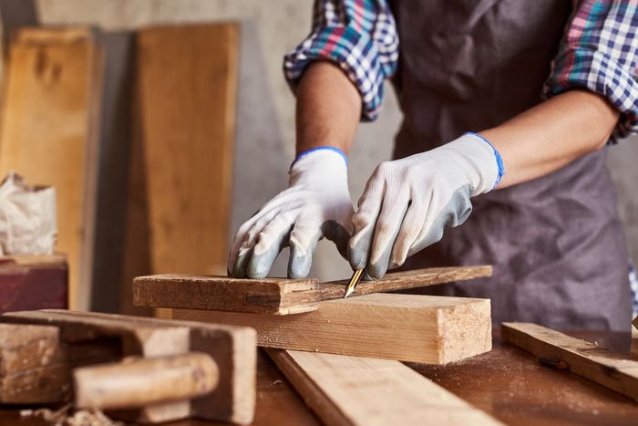 Person in workshop, wearing gloves, marking wood with a pencil on a workbench.