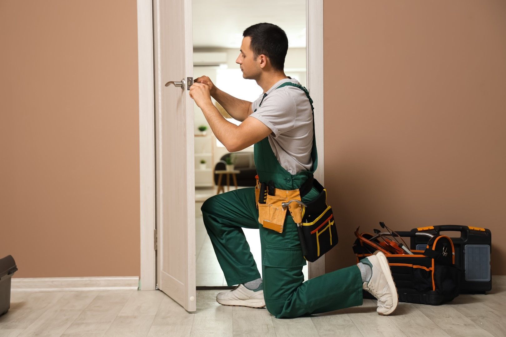 A person kneels to fix a door handle. Wearing green overalls, using a screwdriver, a tool bag is beside.