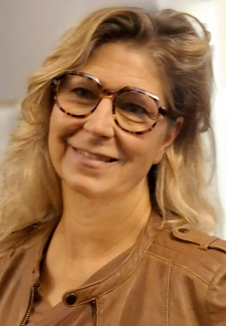 Donna sorridente con occhiali e capelli biondi che indossa una giacca marrone su sfondo chiaro