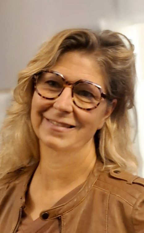 Donna sorridente con occhiali e capelli biondi che indossa una giacca marrone su sfondo chiaro
