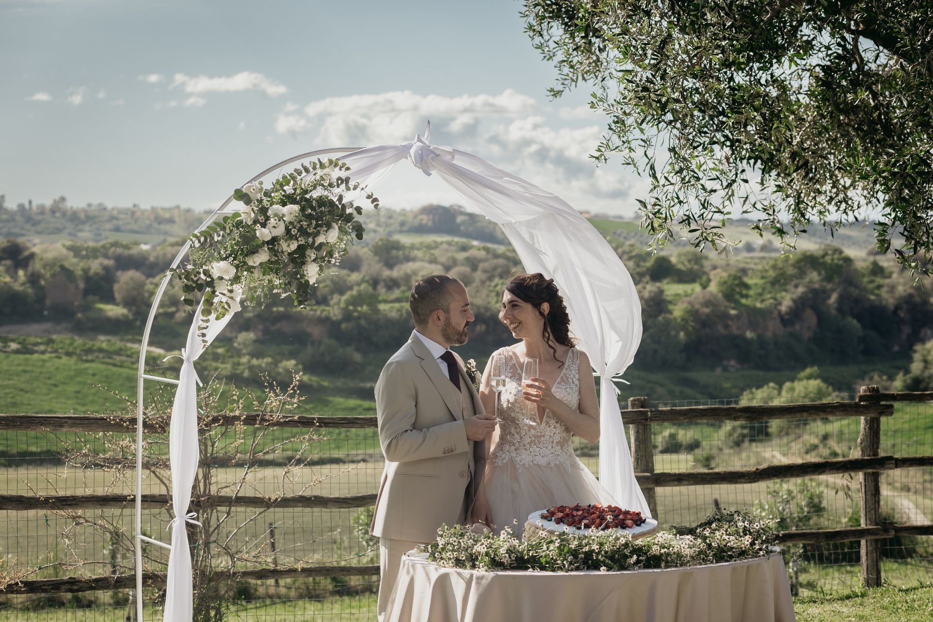 Coppia che brinda con champagne sotto un arco decorato; matrimonio all'aperto con vista panoramica sulla campagna.
