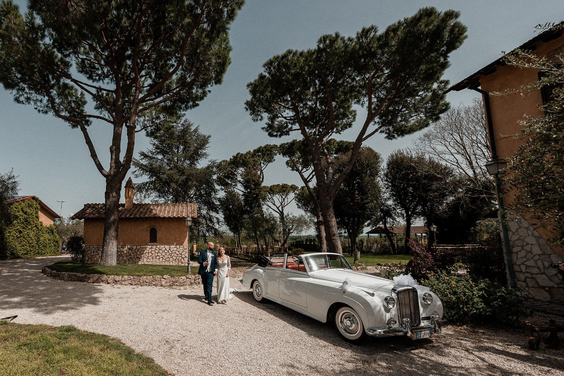 Gli sposi novelli sbarcano da un'auto d'epoca bianca su un vialetto sterrato, tra edifici e alberi.
