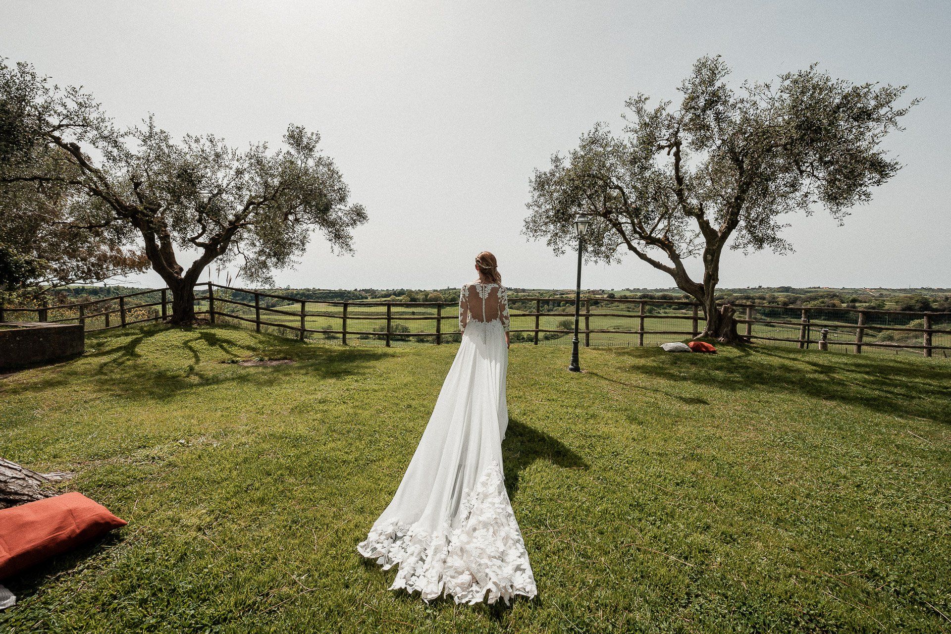 Una sposa con un abito da sposa in pizzo si allontana in un campo erboso verso degli alberi e una recinzione.