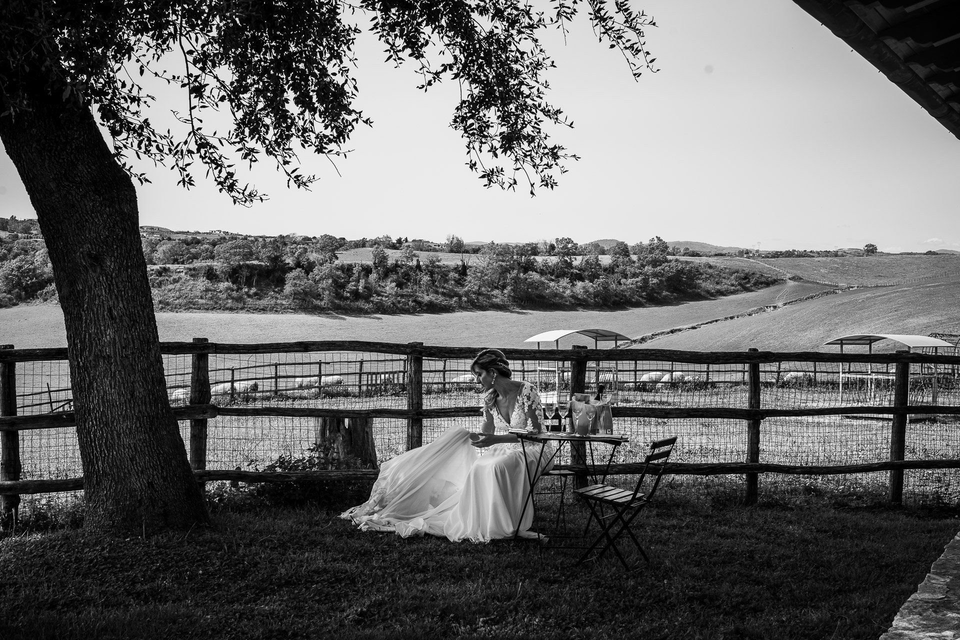 La sposa è seduta a un tavolo, sotto un albero che domina un vigneto, e scrive, in bianco e nero.