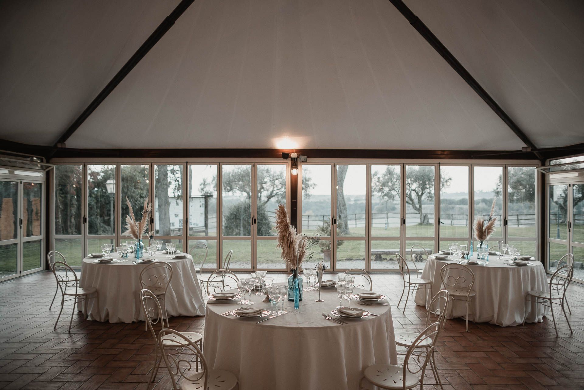 Sala da pranzo formale con tavoli rotondi, tovaglie bianche e pareti in vetro con vista sul paesaggio esterno.