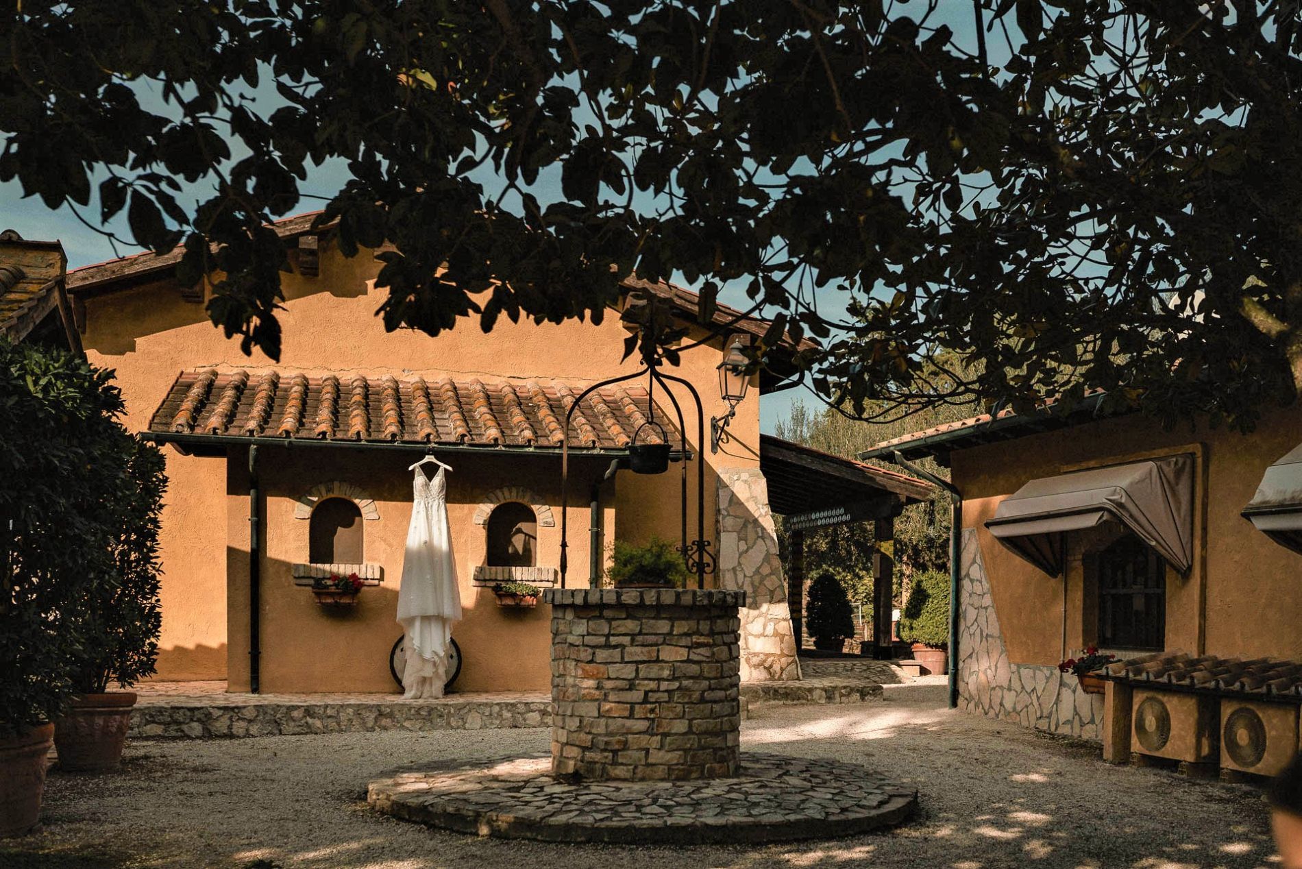 Abito da sposa appeso a un muro vicino a un pozzo in un cortile di edifici in stile italiano, sotto i rami degli alberi.