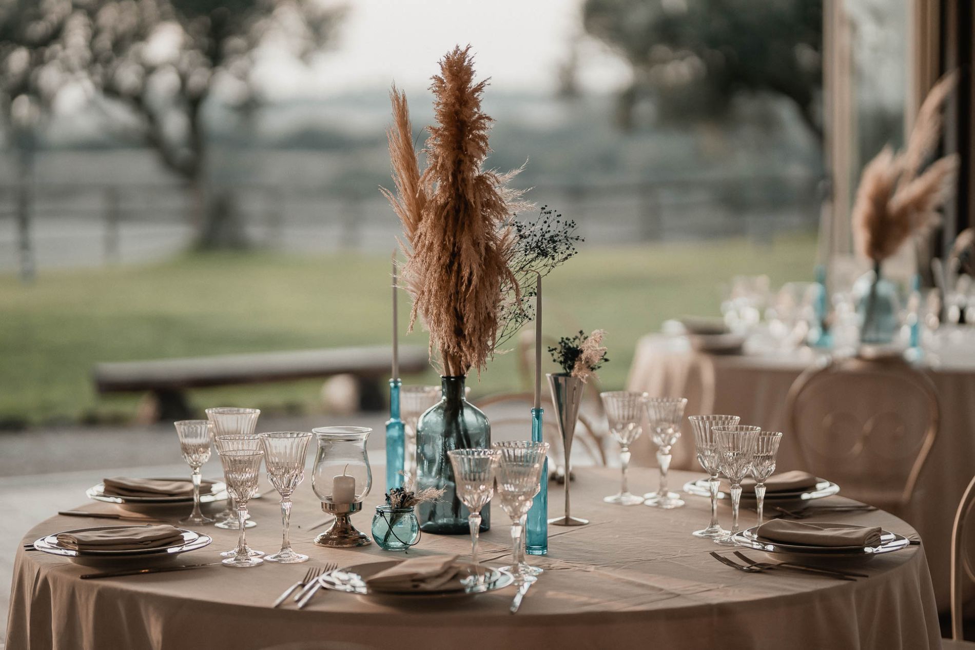 Elegante apparecchiatura da tavola con centrotavola di fiori secchi, vasi blu e bicchieri di cristallo all'aperto.