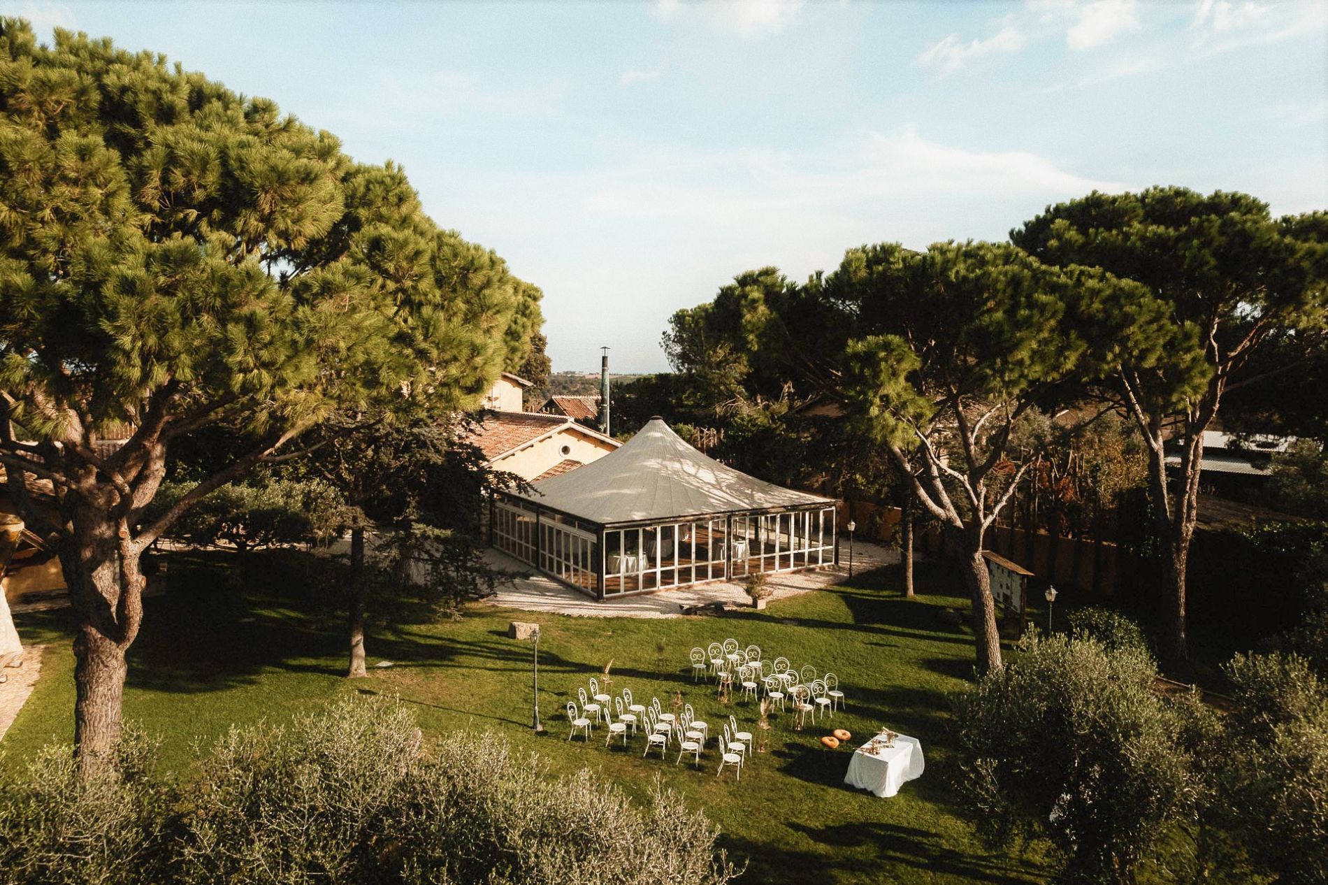 Cerimonia nuziale allestita in giardino, con sedie rivolte verso un gazebo circondato da alberi in una giornata di sole.