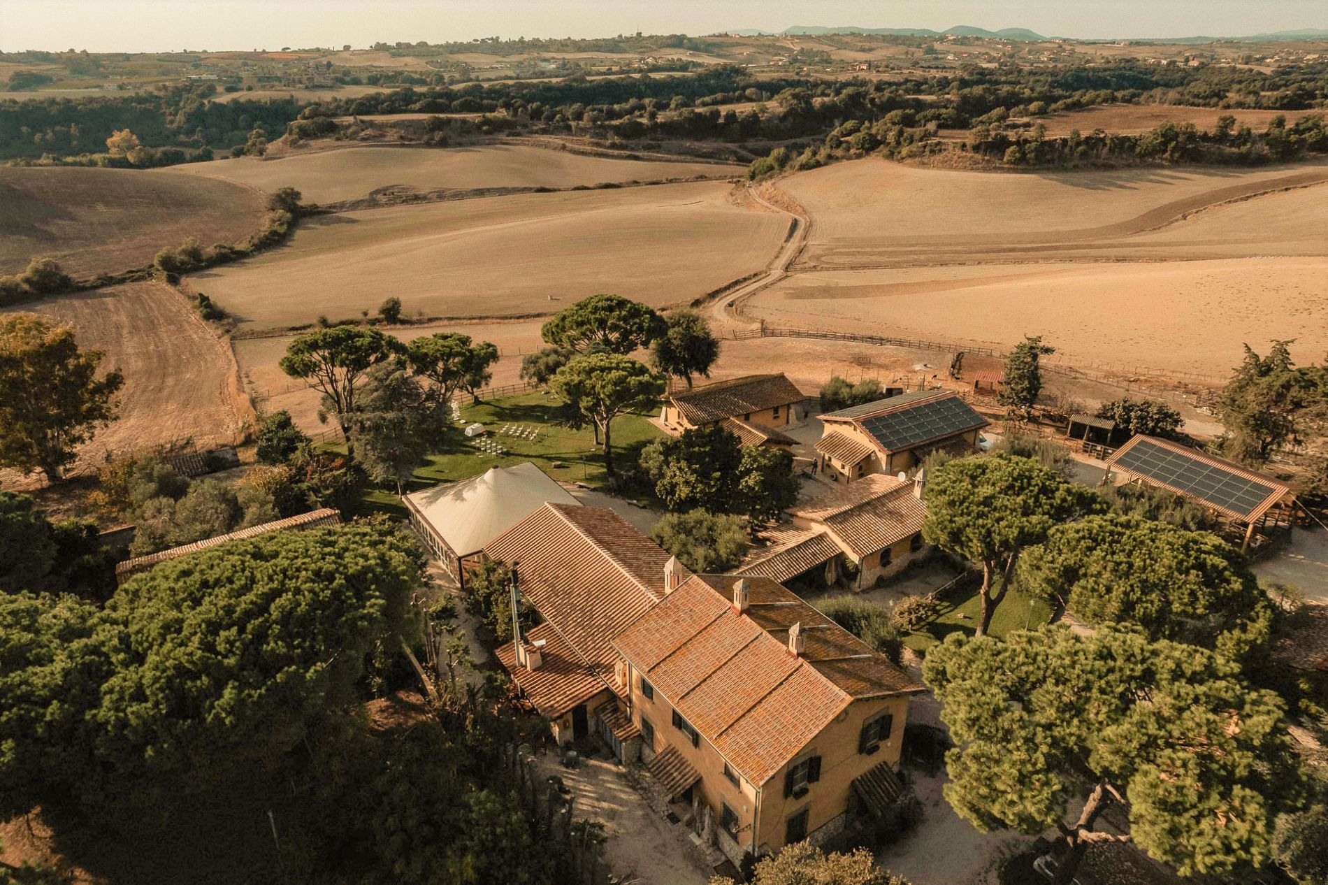 Vista aerea di una fattoria rustica con tetto in terracotta e di edifici circondati da alberi e campi sotto un cielo soleggiato.