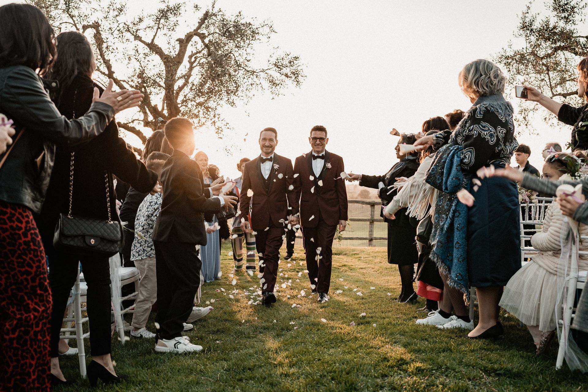 Due sposi camminano, ricoperti di petali di fiori, durante un matrimonio all'aperto. Gli ospiti applaudono, il sole tramonta.