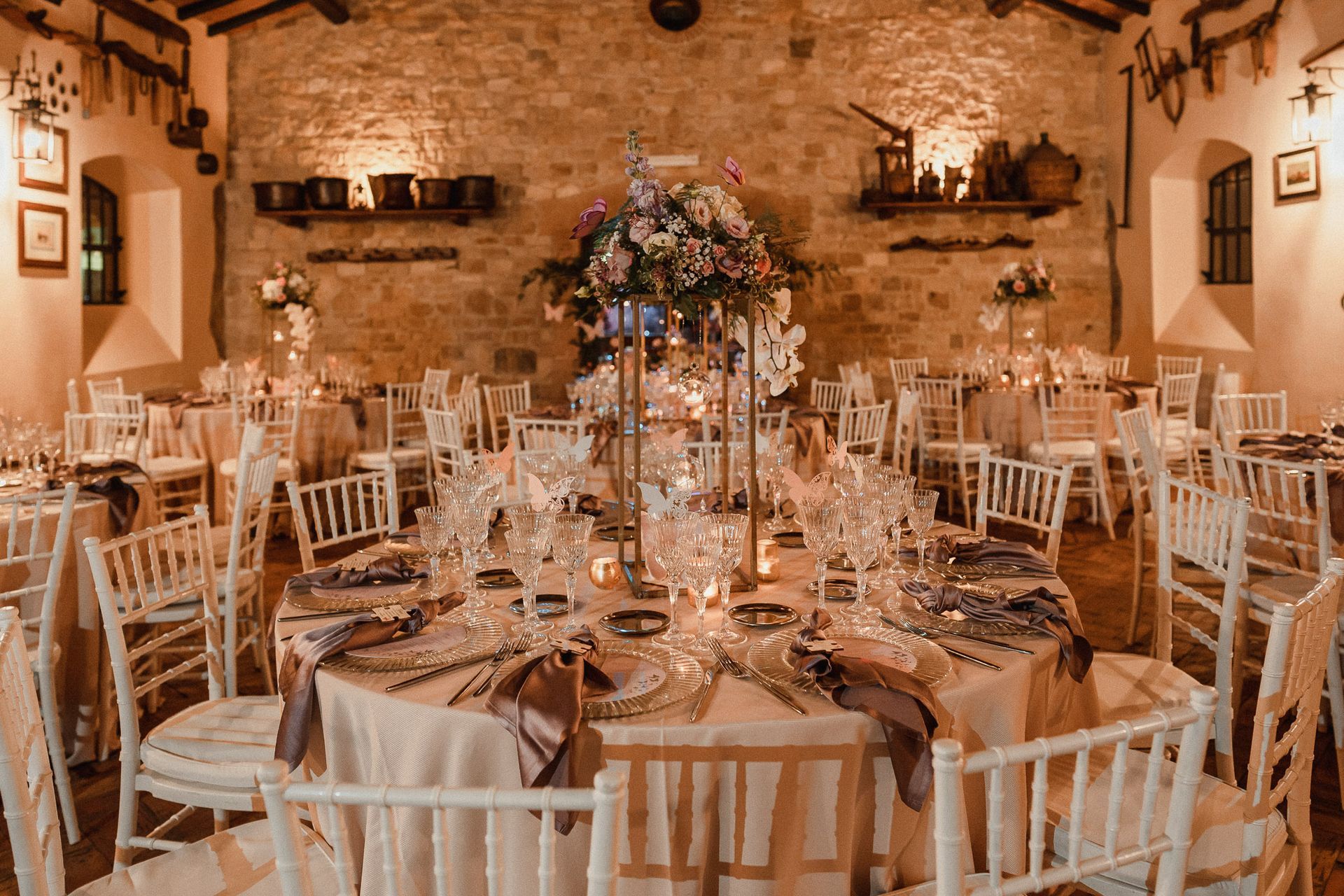 Ricevimento di nozze con tavoli rotondi, fiori e sedie bianche in una sala con pareti in pietra.