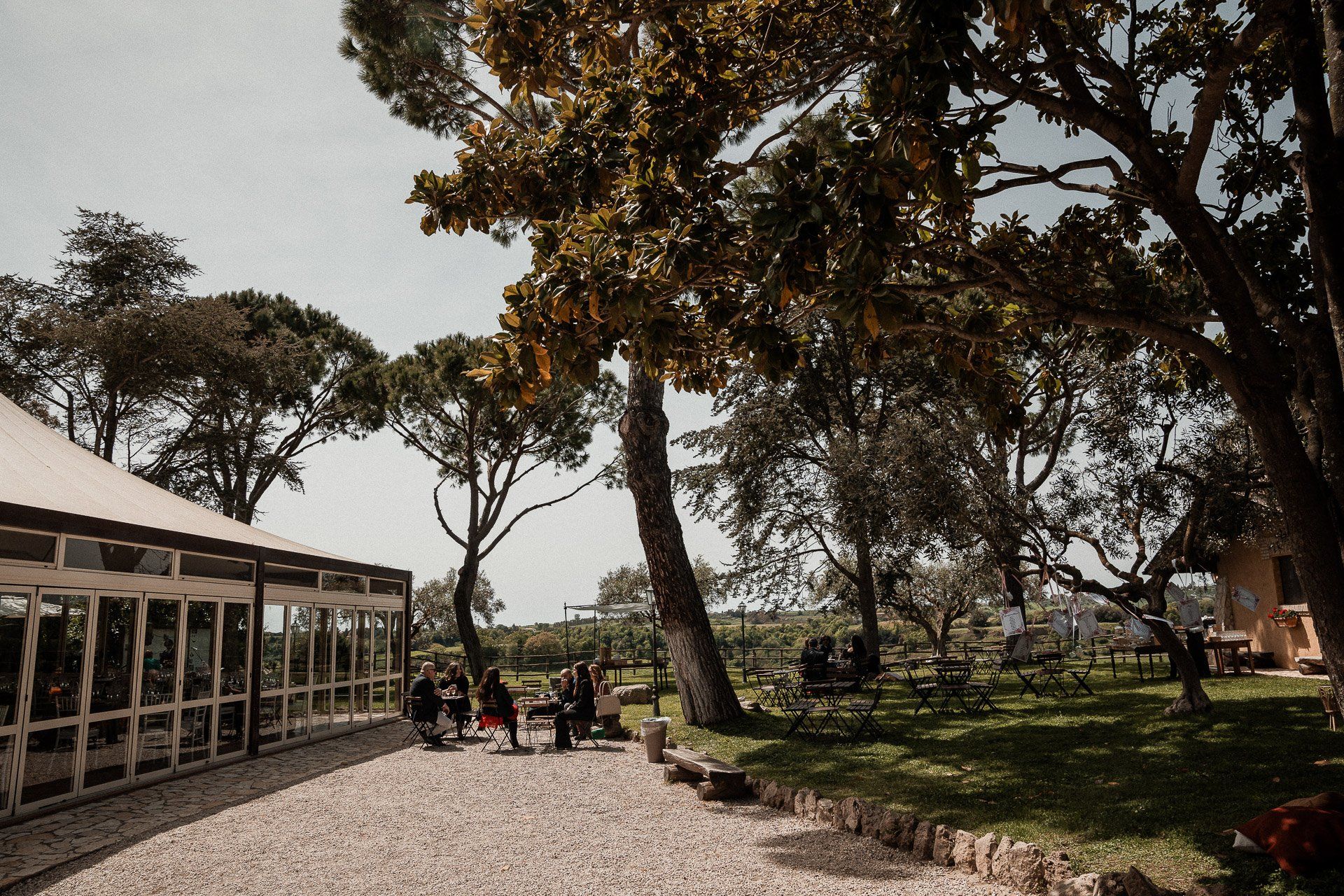 Un sentiero di ghiaia conduce a un edificio con un patio coperto. La gente sosta vicino all'ingresso, circondata da alberi e da un paesaggio soleggiato.