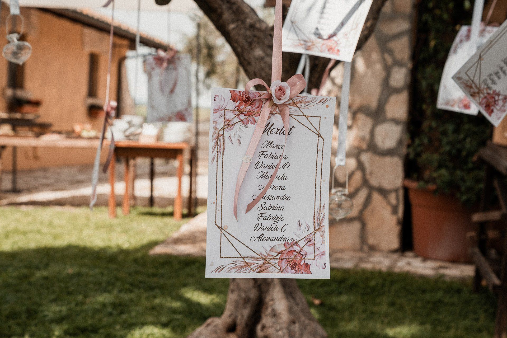 Planimetria per matrimonio appesa a un albero, con decorazione floreale e nastro rosa. Allestimento all'aperto.
