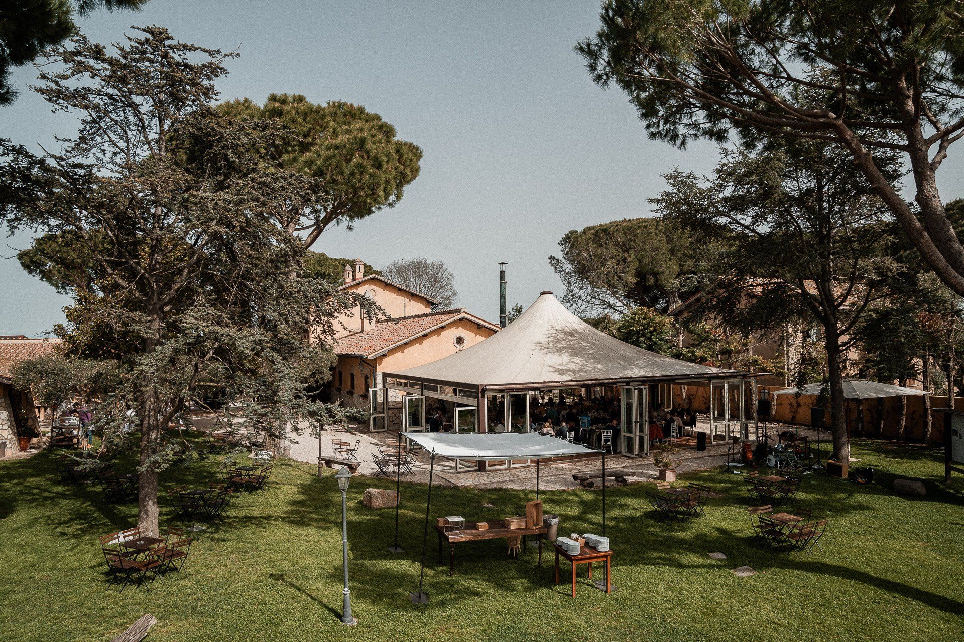 Evento all'aperto: tenda bianca sopra i tavoli, partecipanti in giardino, alberi.