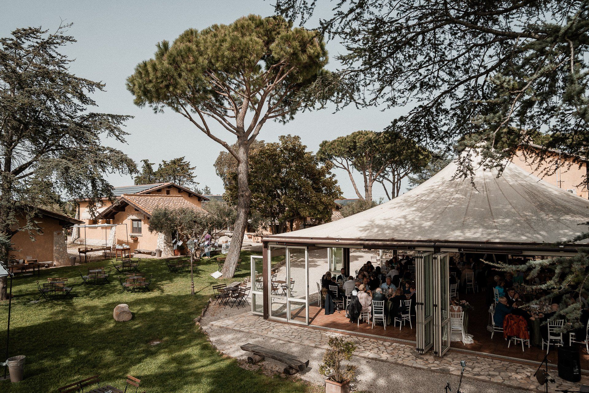 Evento all'aperto sotto una tettoia con persone sedute. Alberi rigogliosi e verdi circondano gli edifici in una giornata di sole.