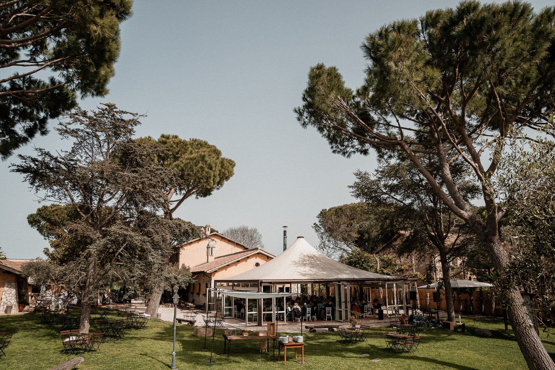 Un prato verde lussureggiante con alberi, un edificio con tavoli e una tenda sullo sfondo, cielo azzurro.