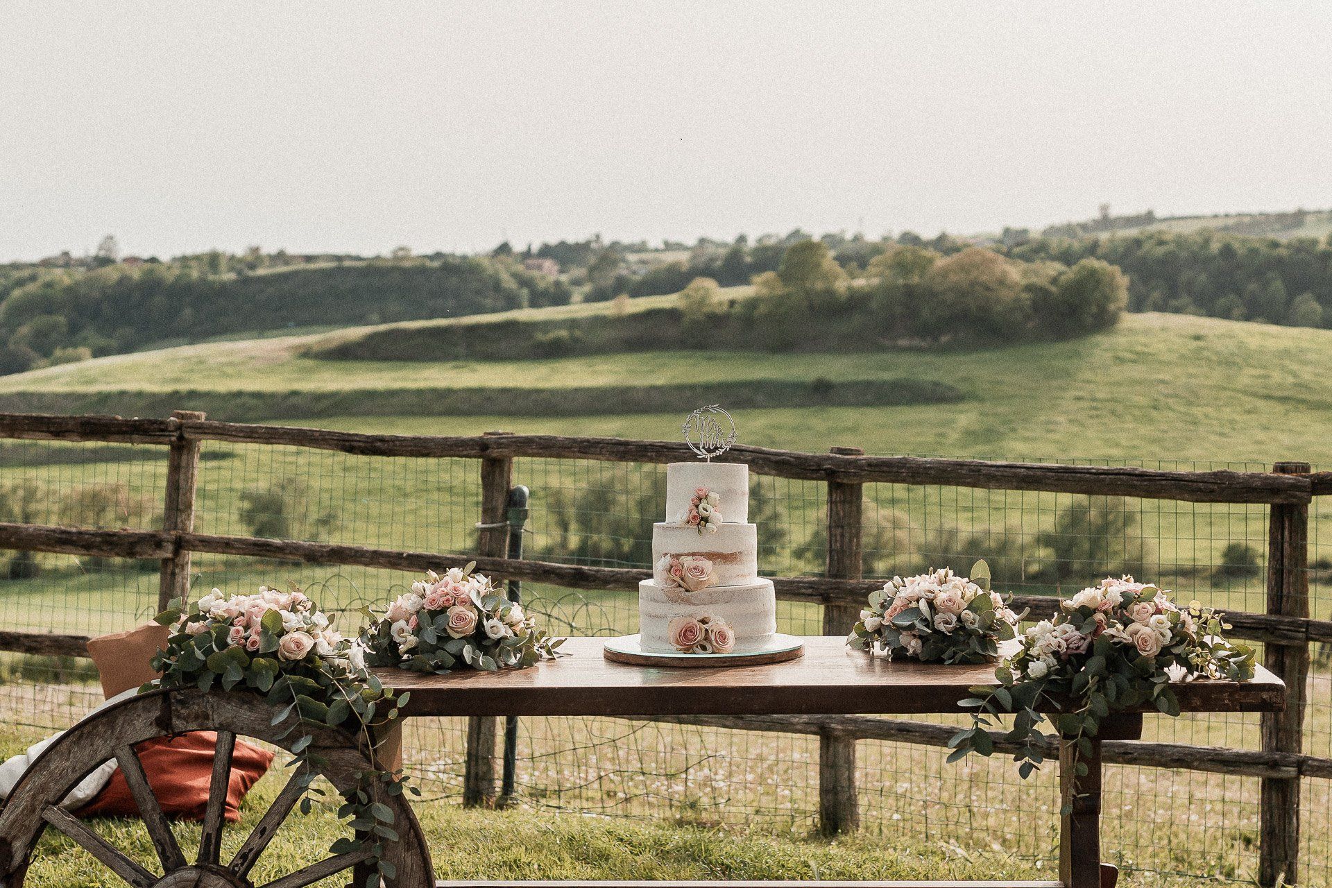 Torta nuziale su un tavolo rustico decorato con fiori; sullo sfondo dolci colline verdi.