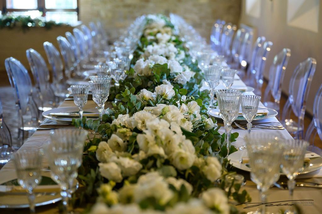 Lungo tavolo apparecchiato per un evento formale, con fiori bianchi, verde e sedie trasparenti.