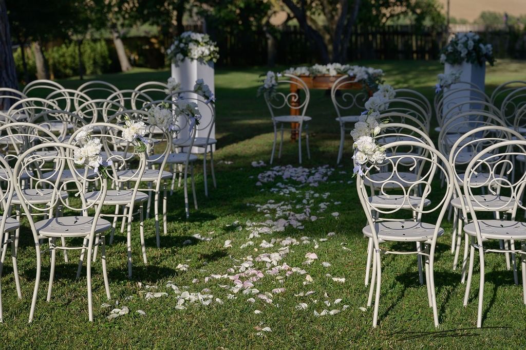 Allestimento per la cerimonia nuziale: sedie bianche, petali di fiori, altare decorato su prato verde.