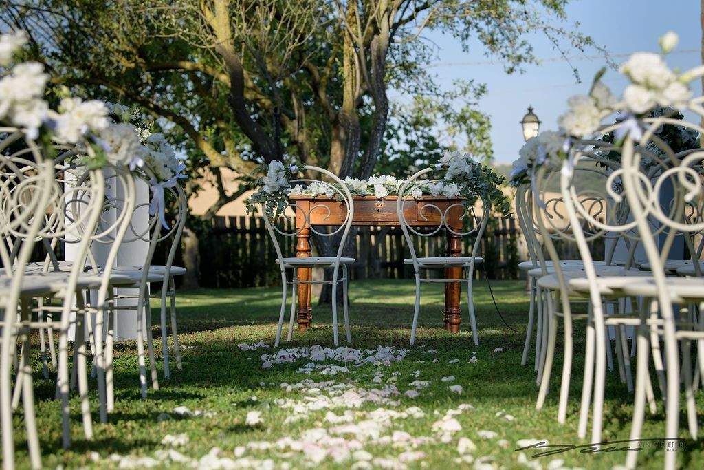 Allestimento della cerimonia nuziale: sedie bianche, altare decorato con fiori e petali sull'erba in un ambiente all'aperto.