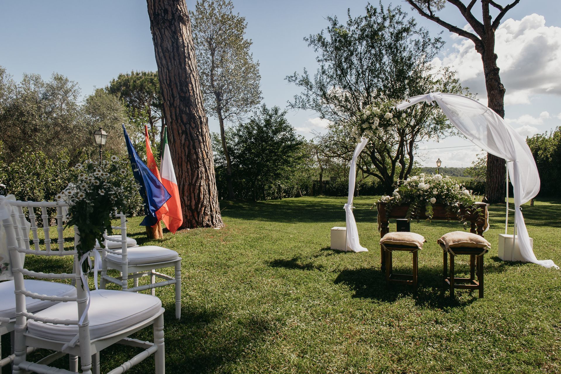 Allestimento per la cerimonia nuziale in un giardino soleggiato: sedie, arco floreale, bandiere e verde.
