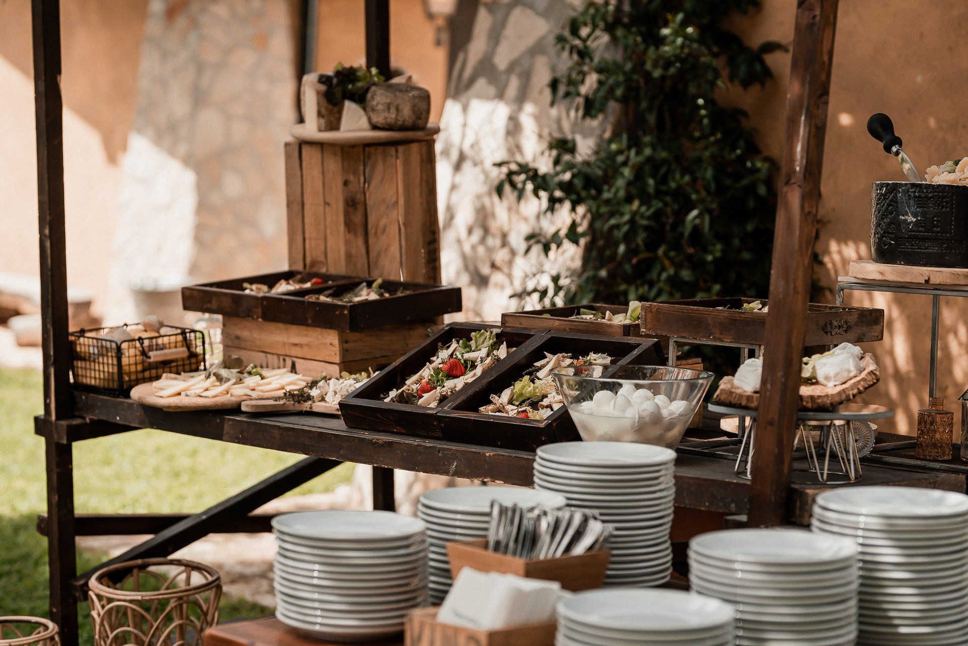 Tavolo da buffet all'aperto con vassoi di cibo, pile di piatti bianchi e una torta scura.