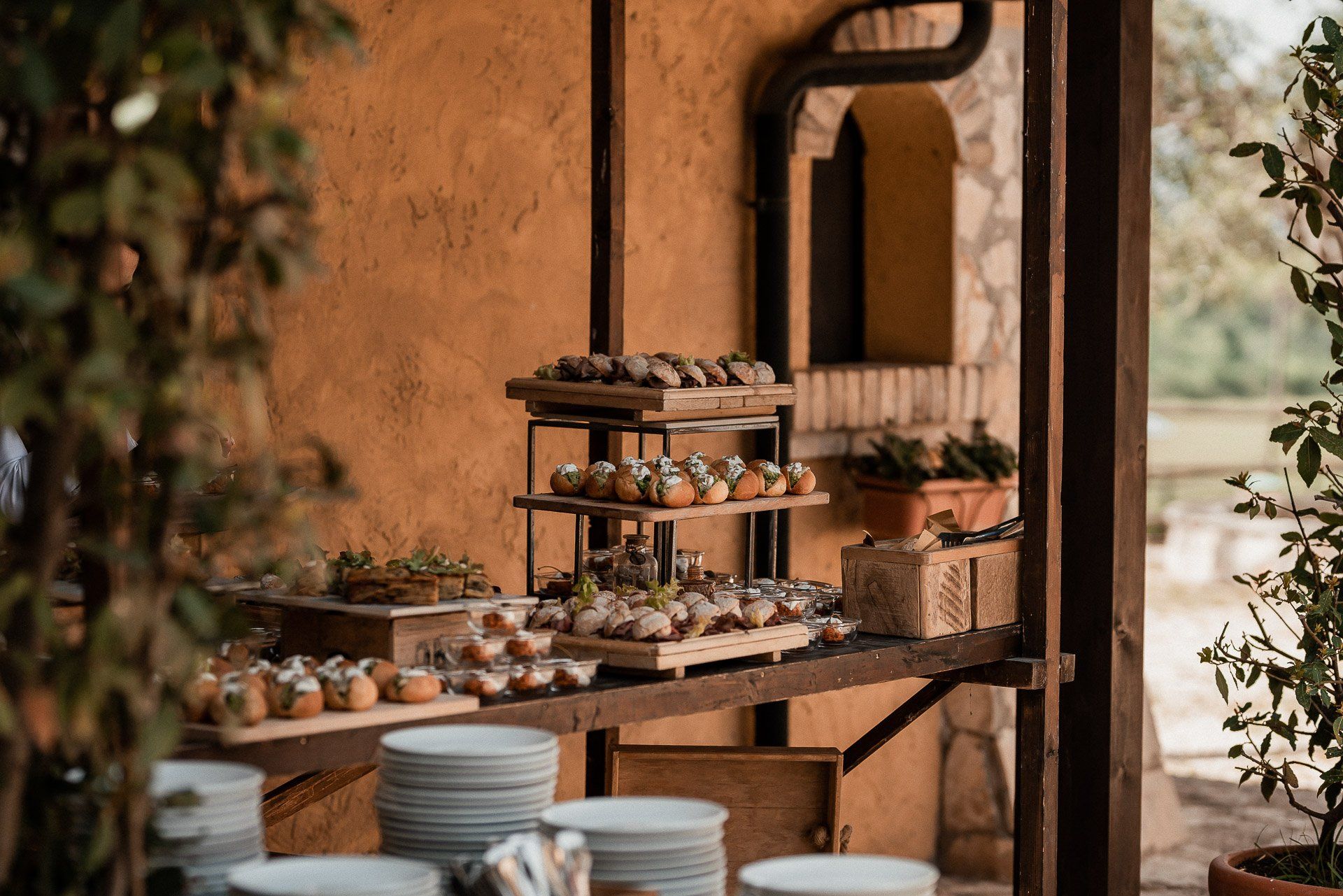 Buffet con pasticcini impilati su piattaforme di legno, piatti bianchi e piante, all'aperto vicino a un edificio rustico.