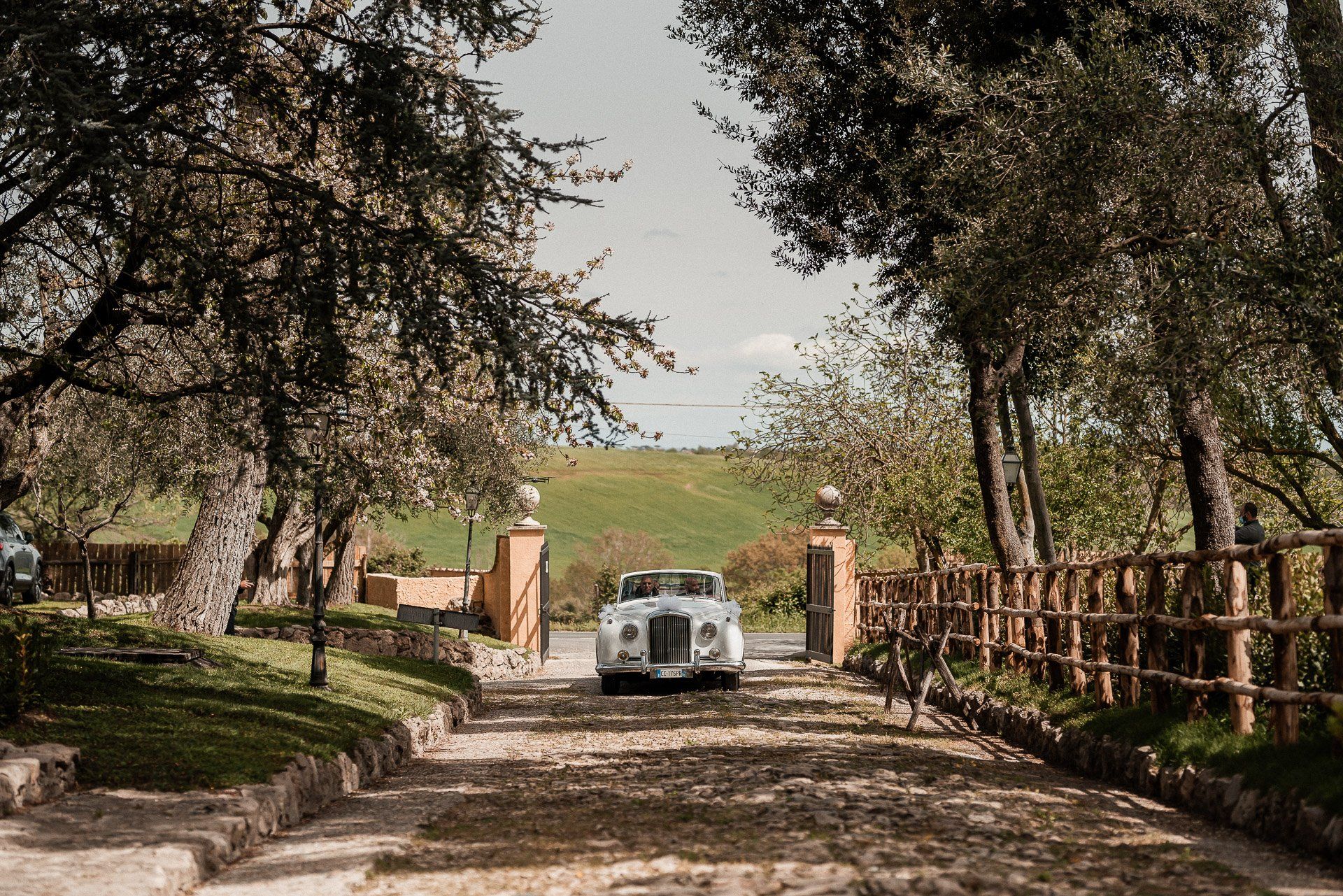 Un'auto d'epoca bianca parcheggiata davanti al cancello d'ingresso di una villa; sullo sfondo si vedono dolci colline.