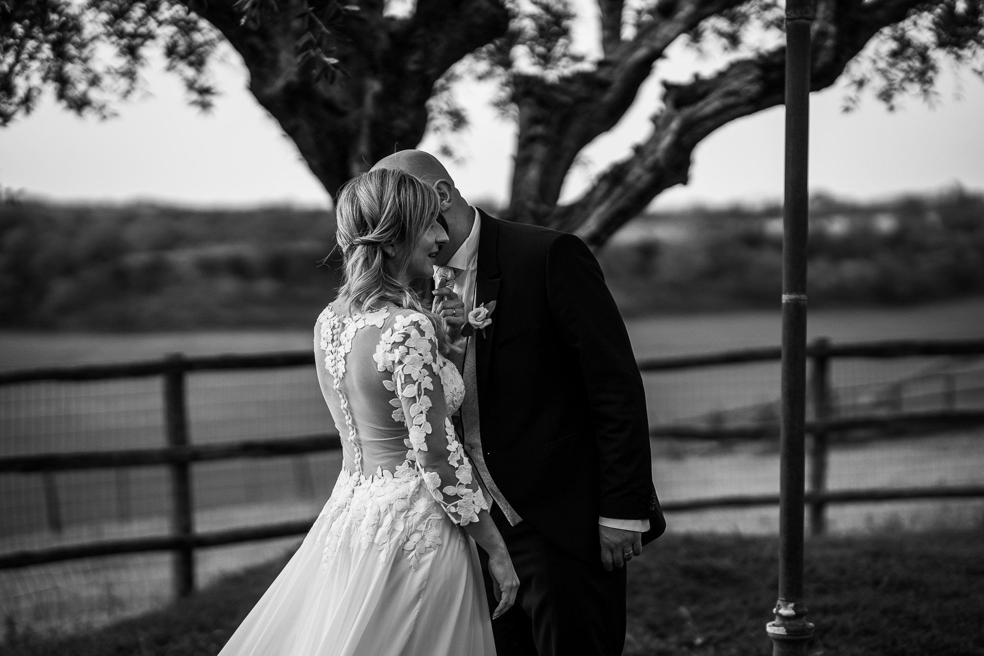 Gli sposi condividono un momento di tenerezza. Lo sposo bacia la guancia della sposa; sono all'aperto, vicino a un albero.