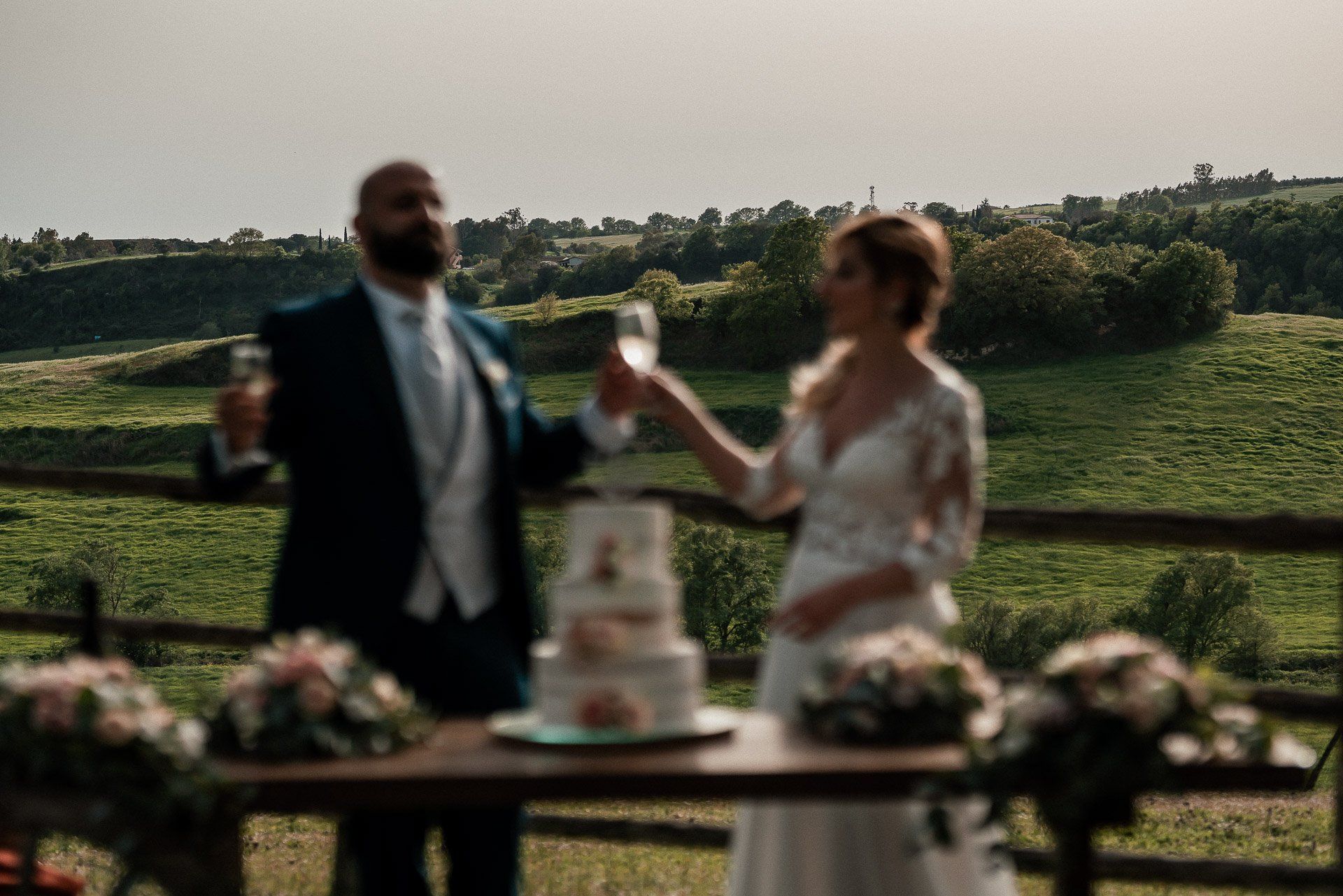 Coppia che brinda con calici di champagne a un matrimonio. Torta sul tavolo con fiori; sfondo panoramico all'aperto.