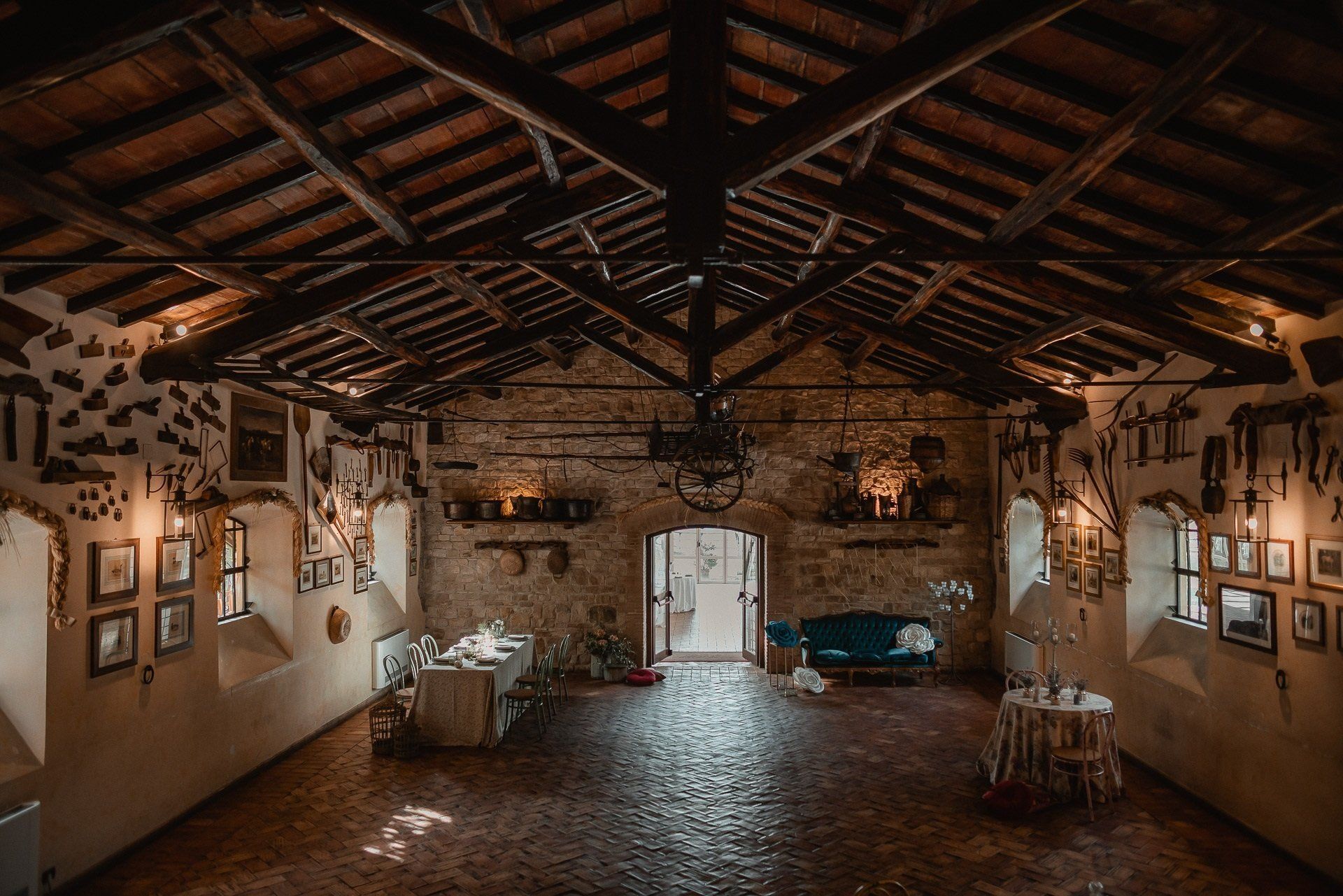 Interno rustico di un edificio in pietra con travi in legno, caratterizzato da tavoli, un divano blu e opere d'arte incorniciate.