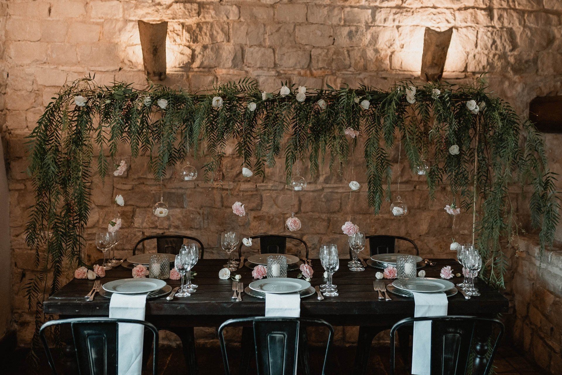 Apparecchiatura per il tavolo di nozze con sfondo verde, fiori rosa e sedie nere in una stanza con pareti in pietra.