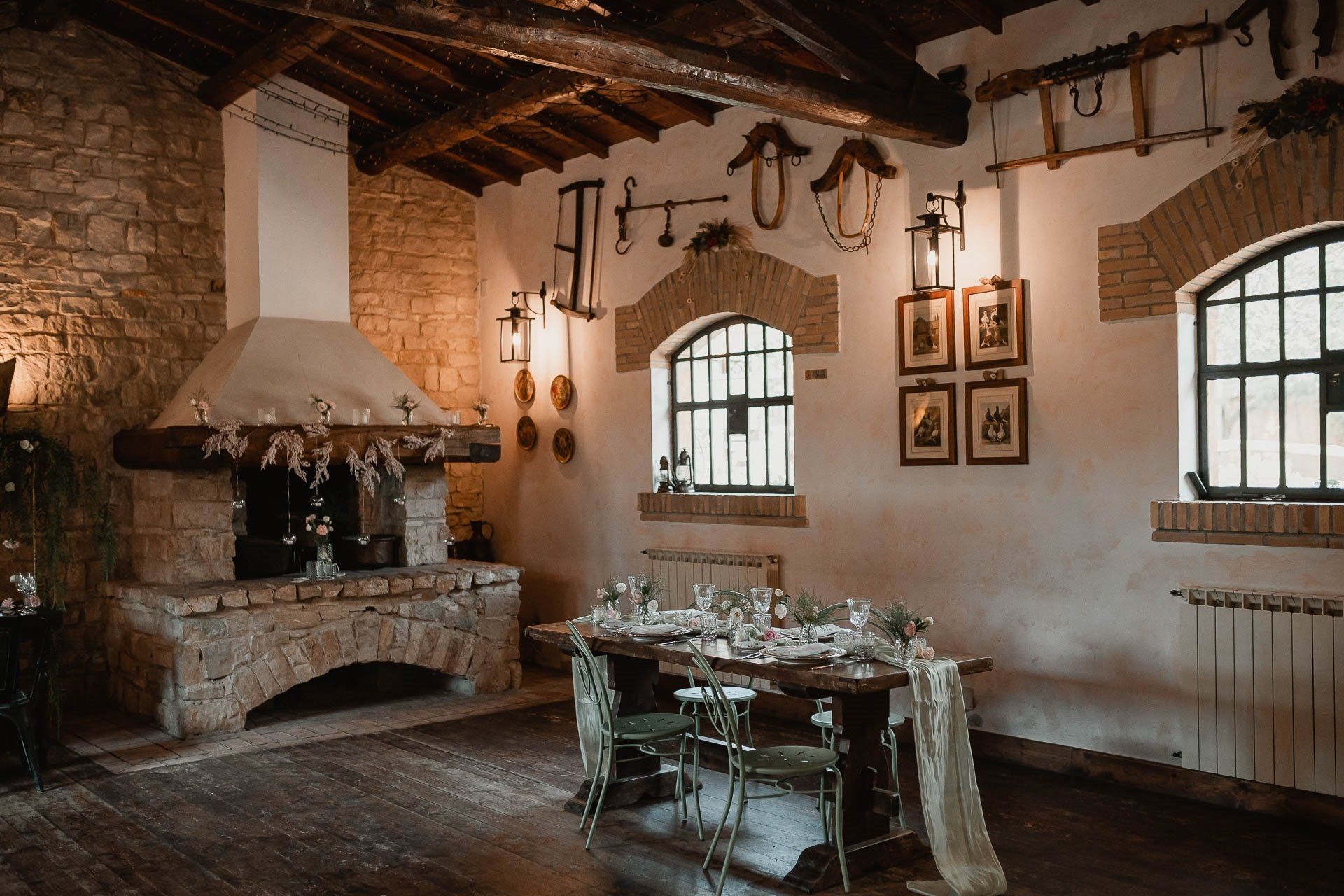 Sala da pranzo rustica con camino in mattoni, tavolo apparecchiato e finestre ad arco.