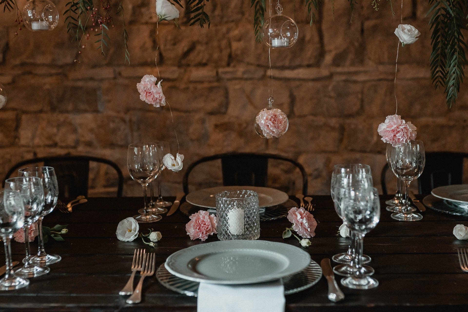 Apparecchiatura della tavola: tavolo in legno scuro, apparecchiati, decorazioni floreali, sfere di vetro appese, contro un muro di pietra.