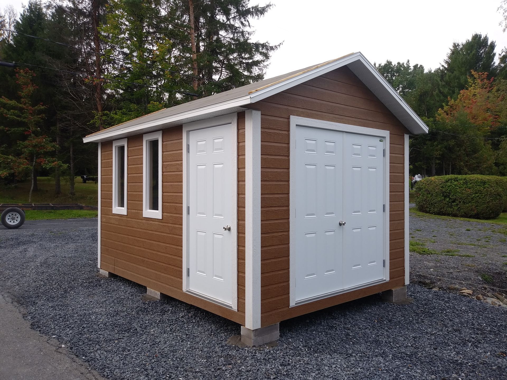 Abri de jardin marron et blanc avec porte de garage, deux fenêtres et une porte sur un sol en gravier.