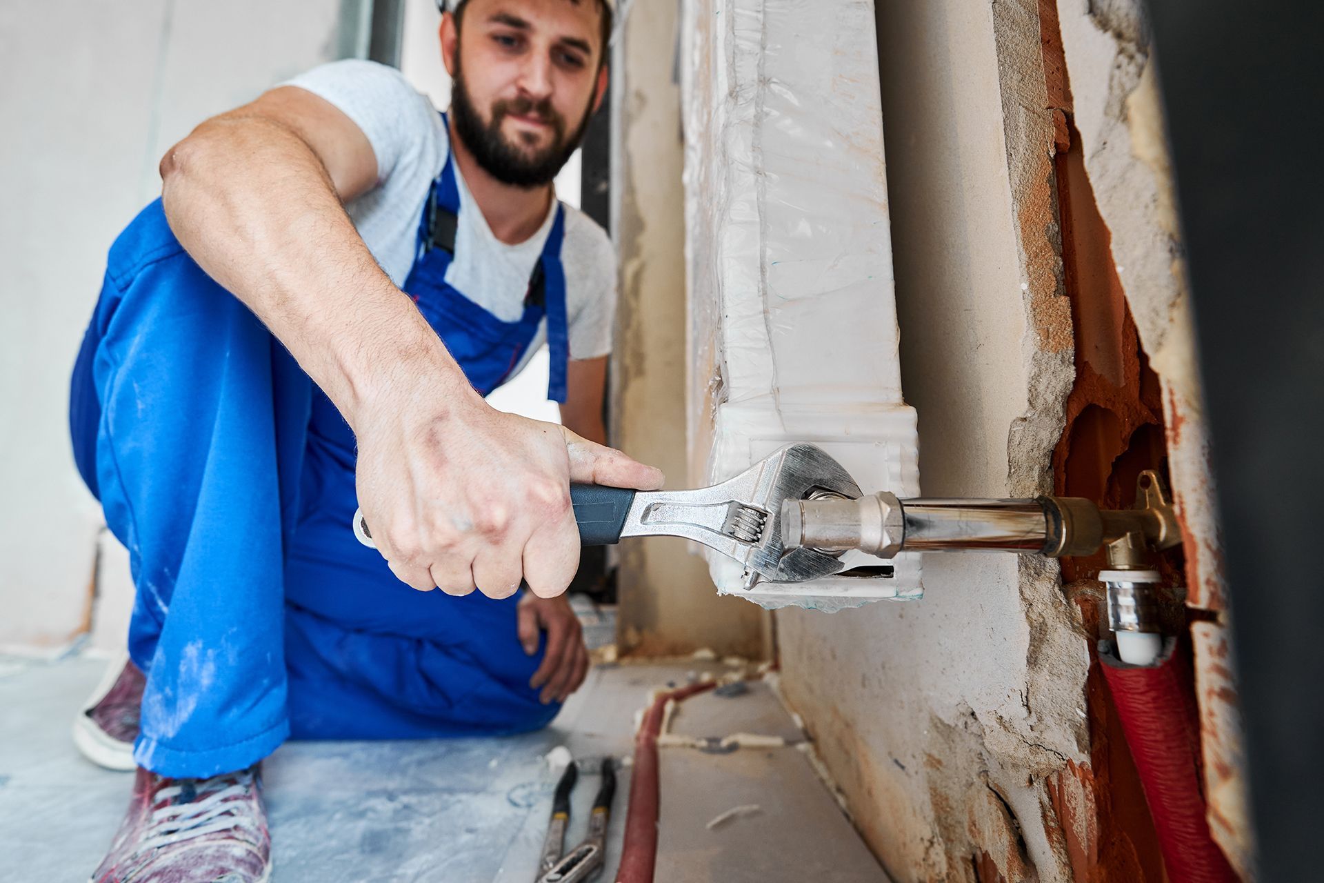Plumber in blue overalls, using a wrench on a pipe in a wall.