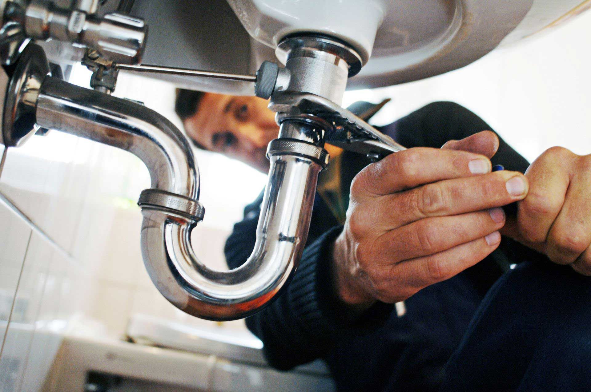 A plumber from a plumbing company, in a blue uniform, repairing the sink drain with a wrench. Plumber working under a sink with a wrench. Chrome pipes and white sink in a bathroom setting.