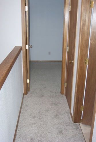 A bathroom with a toilet , sink and vacuum cleaner. Hallway with carpet, two wooden doors, and a doorway leading to a room with bare walls.