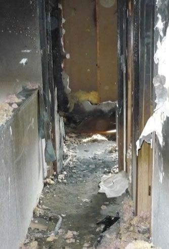 A bathroom with a toilet and a wooden stool. Hallway interior severely damaged by fire; charred walls, debris on floor.