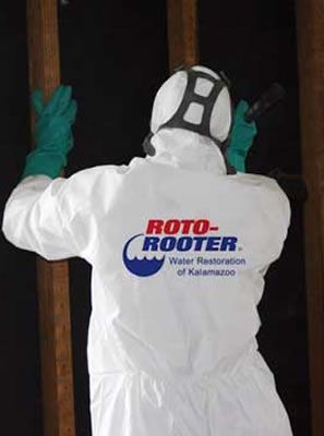 A man wearing a white protective suit and gloves is working on a wall. Person in white protective suit from Roto-Rooter, inspecting structure with flashlight. Green gloves, black background.