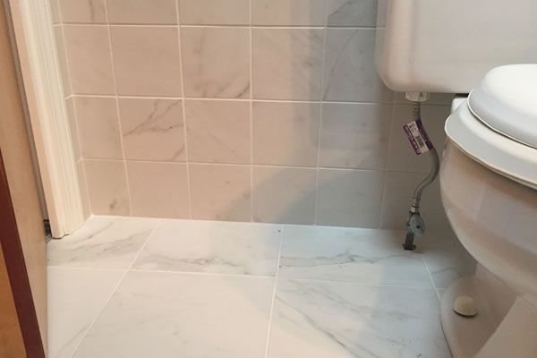 A toilet is sitting in a bathroom next to a door. Bathroom with white marble tile floor and walls, toilet on the right.