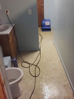 A bathroom with a toilet , sink and vacuum cleaner. Bathroom with wet floor, fan, sink, toilet, and tangled power cord.