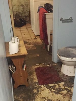 A bathroom with a toilet and a wooden stool. Bathroom with soiled floor, toilet, small table with toilet paper. A washing machine is visible in the background.